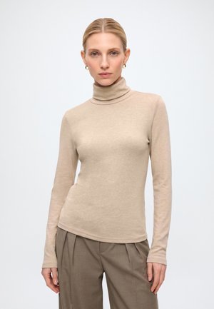 Femme aux cheveux blonds portant un pull à col roulé beige et un pantalon plissé marron sur un fond uni clair.