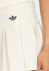 Biela plisovaná tenisová sukňa z textúrovanej tkaniny, s čiernym logom Adidas na páse. Minimalistický dizajn, hladký finiš.
