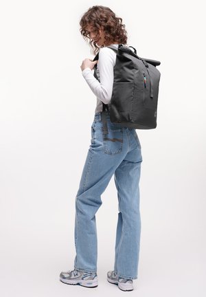 Persona de cabello rizado con camisa blanca de manga larga, jeans azul claro, zapatillas grises, llevando una mochila negra grande con cierre enrollable.