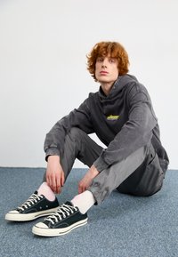 Haut à capuche gris avec logo, pantalon gris fuselé, et baskets en toile vert foncé avec bout blanc et lacets blancs. Chaussettes courtes roses claires visibles.