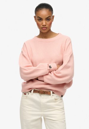 Roze gebreide trui met een brede halslijn en oversized mouwen, gecombineerd met witte broek en een gevlochten bruine riem. Gestructureerde, zachte stof.