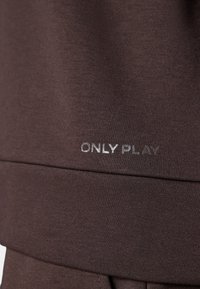 Кафява тъкан с близък план на шев и думите ONLY PLAY, отпечатани с малки, светлосиви букви близо до шева.