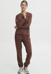 Felpa e pantaloni jogger marroni, realizzati in tessuto morbido; presentano una vestibilità comoda, polsini a coste e cuciture accentuate. Abbinati a sneakers chiare.