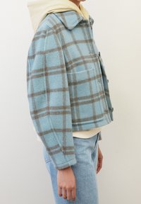 Giacca a quadri blu realizzata in un materiale morbido simile alla lana, con un design a collare, una tasca frontale e un taglio corto sopra un leggero hoodie.