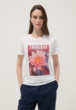 Jeune femme portant un t-shirt blanc avec une grande fleur de lotus rose et le texte « THAILAND », associée à un pantalon bleu foncé, debout les mains dans les poches.