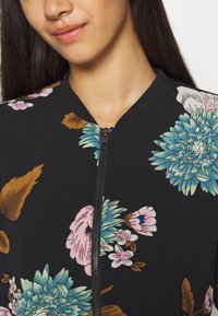 Zwarte bomberjack met bloemenprint, uitgevoerd in levendige blauw-, roze- en goudtinten, voorzien van een rits en geribde kraag. Soepele textuur.