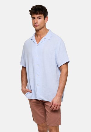 Hombre vistiendo una camisa de manga corta de color azul claro abotonada y pantalones cortos marrones, de pie con una mano en el bolsillo.