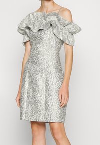 Femme portant une robe argentée texturée longueur genou avec des manches volantées épaules dénudées et une taille cintrée sur un fond uni.