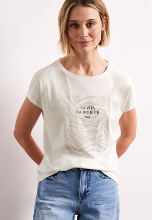Femme aux cheveux blonds courts portant un t-shirt blanc avec le texte "LA VITA DA BOHÈME Italy" et un jean bleu clair, debout les mains derrière le dos.