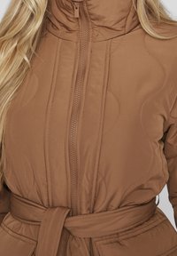 Veste matelassée marron avec un col montant, une fermeture éclair frontale et une ceinture nouée. Présente des coutures courbes et une texture lisse et brillante.