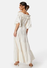 Lang hvid off-shoulder kjole med flæser, korte pufærmer, taljebælte og lagdelt nederdel i et textureret stof.