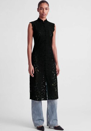 Femme portant un long gilet en dentelle noire sans manches avec col mandarin sur un jean bleu clair et des chaussures noires pointues sur fond blanc.