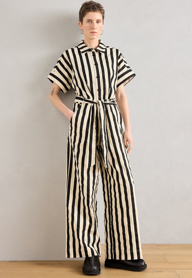 Μονόχρωμη ολόσωμη φόρμα με μαύρες και κρεμ ρίγες, κοντά μανίκια, κουμπιά μπροστά, ζώνη στη μέση και φαρδιά πόδια. Υφασμάτινη υφή και τσέπες στα πλάγια.