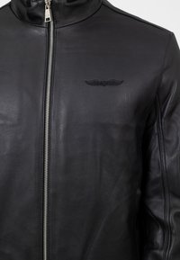 Giacca in pelle nera con colletto alto, zip frontale e ricamo di ali sul lato sinistro del petto. Texture liscia e design aderente.