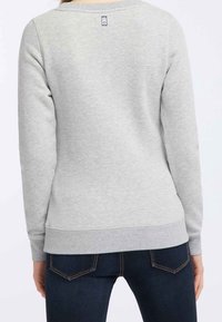 Grijze sweatshirt met een geribbelde nekline en manchetten, voorzien van een klein logo-label op de bovenrug. Draagt met donkere jeans.