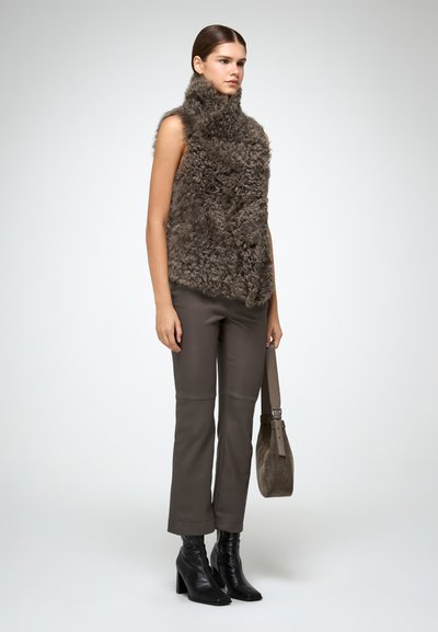 Gilet in finta pelliccia marrone con colletto alto, indossato su pantaloni attillati marrone scuro e stivali neri con tacco, mentre tiene una borsa a tracolla grigia.
