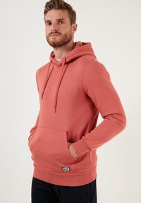 Sudadera con capucha color coral que presenta un bolsillo frontal, puños y dobladillo acanalados, confeccionada con una tela suave y de textura lisa.
