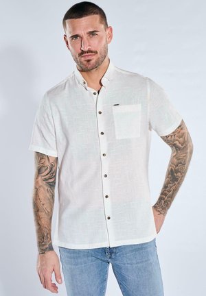 Man with short hair and tattoos on both arms wears a white short-sleeve button-up shirt and light blue jeans, standing with one hand in pocket.