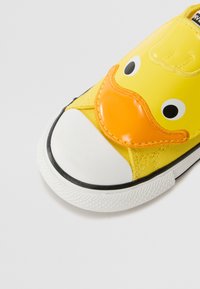 Kollane kanvasking, millel on valge kummist varbaosa, millel on parte näo disain suurte silmade ja oranži nokaga, tekstuuriga õmbluste detailidega.
