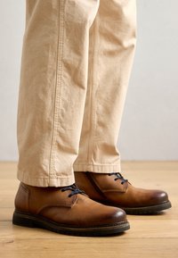 Brown leather shoes with rounded toes, black soles, and blue laces; paired with light beige pants resting on a wooden floor.