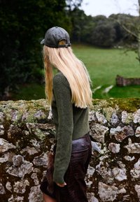 Oseba z dolgimi blond lasmi, ki nosi temno kapo, zeleno pulover in temno krilo, stoji ob kamnitem zidu, pokritem z mahom, in gleda preko zelenega polja.