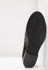 Chaussure en cuir verni noir avec un bout arrondi, des accents de brogue détaillés et une semelle en caoutchouc noir texturé, positionnée sur un fond blanc.