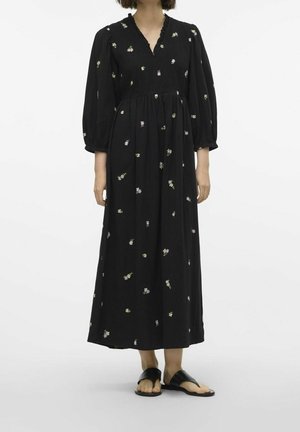 Robe maxi noire à manches longues avec petits imprimés floraux, taille froncée, décolleté en V avec volants, portée avec des sandales plates noires ouvertes au bout.