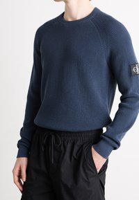 Pull en maille bleu marine avec un motif texturé, col ras du cou et poignets côtelés. Présente une étiquette de logo sur la manche gauche. Associé à un short noir.