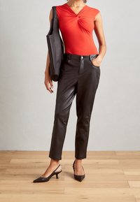 Osoba nosząca czerwoną bluzkę bez rękawów z skręconym przodem, czarne skórzane spodnie, czarne sandały na obcasie, oraz trzymająca czarną torbę na ramię.