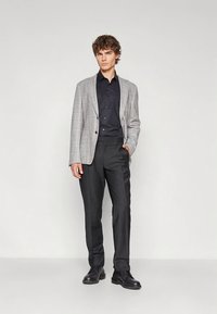 Grijze geruite blazer over een zwart shirt, gecombineerd met zwarte broek en zwarte schoenen. De blazer heeft bruine knopen en een getailleerde pasvorm.