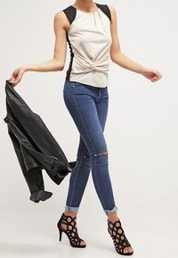 Off-white ärmlös topp med vridningsdetalj, matchad med mörkblå slitna skinny jeans och svarta öppna ankelboots med cirkulära utskärningar.