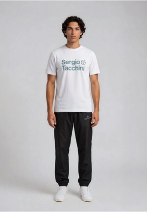 Ung mand står vendt fremad iført hvid Sergio Tacchini T-shirt, sorte joggers og hvide sneakers mod en ensartet lysegrå baggrund.
