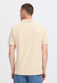 Chemise polo beige en tissu texturé, dotée d'un col classique et de manches courtes, présentée de dos avec une coupe droite.