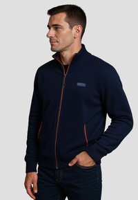 Marine blauwe ritstrui met hoge kraag, oranje rits en zakaccenten, gepresenteerd op een model tegen een neutrale achtergrond.