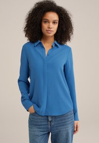 Camicia blu a maniche lunghe con colletto button-down, tessuto strutturato e vestibilità morbida, abbinata a jeans blu chiaro.