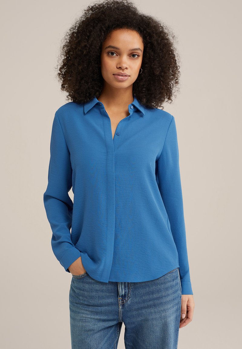 Camicia blu a maniche lunghe con colletto button-down, tessuto strutturato e vestibilità morbida, abbinata a jeans blu chiaro.