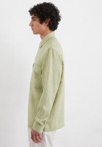Chemise à manches longues vert clair en lin, avec un col boutonné et une poche sur la poitrine. Vue latérale, texture lisse, coupe ample.