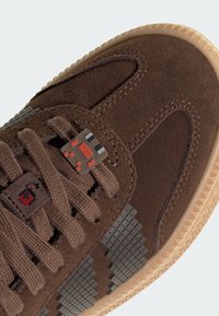 Braune Wildleder-Sneaker mit grauen pixelierten Akzenten, strukturiertem Nähen und einem roten und grauen Dekorationsanhänger an den Schnürsenkeln. Gummisohle.