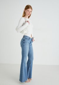 Mujer que lleva una blusa blanca acanalada con botones, jeans acampanados de cintura alta en un tono azul claro, sandalias blancas y un bolso blanco de hombro, de pie de lado.