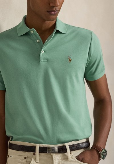 Moški v svetlo zeleni polo majici z majhnim izvezenim logotipom konja, bež hlačah, temnem pasu in ročni uri z rjavim usnjenim paščkom.