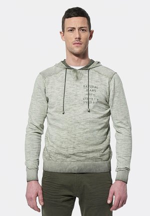 Sudadera verde claro con un acabado texturizado, cordones y un gráfico en la parte delantera. Puños y dobladillo acanalados, llevada encima de pantalones verde oliva.
