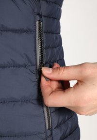 Mörkblå pufferväst med quiltad textur, som har en sidoknapp. Hand visas hållande i dragkedjan, som har en grå accent.