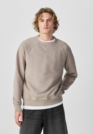 Young man with wavy blonde hair wearing a beige sweatshirt over a white shirt and dark jeans, standing against a plain light background.