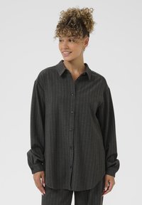 Donna con capelli ricci, che indossa una camicia button-up grigio scuro a righe e pantaloni abbinati, sorridente e con lo sguardo rivolto di lato.