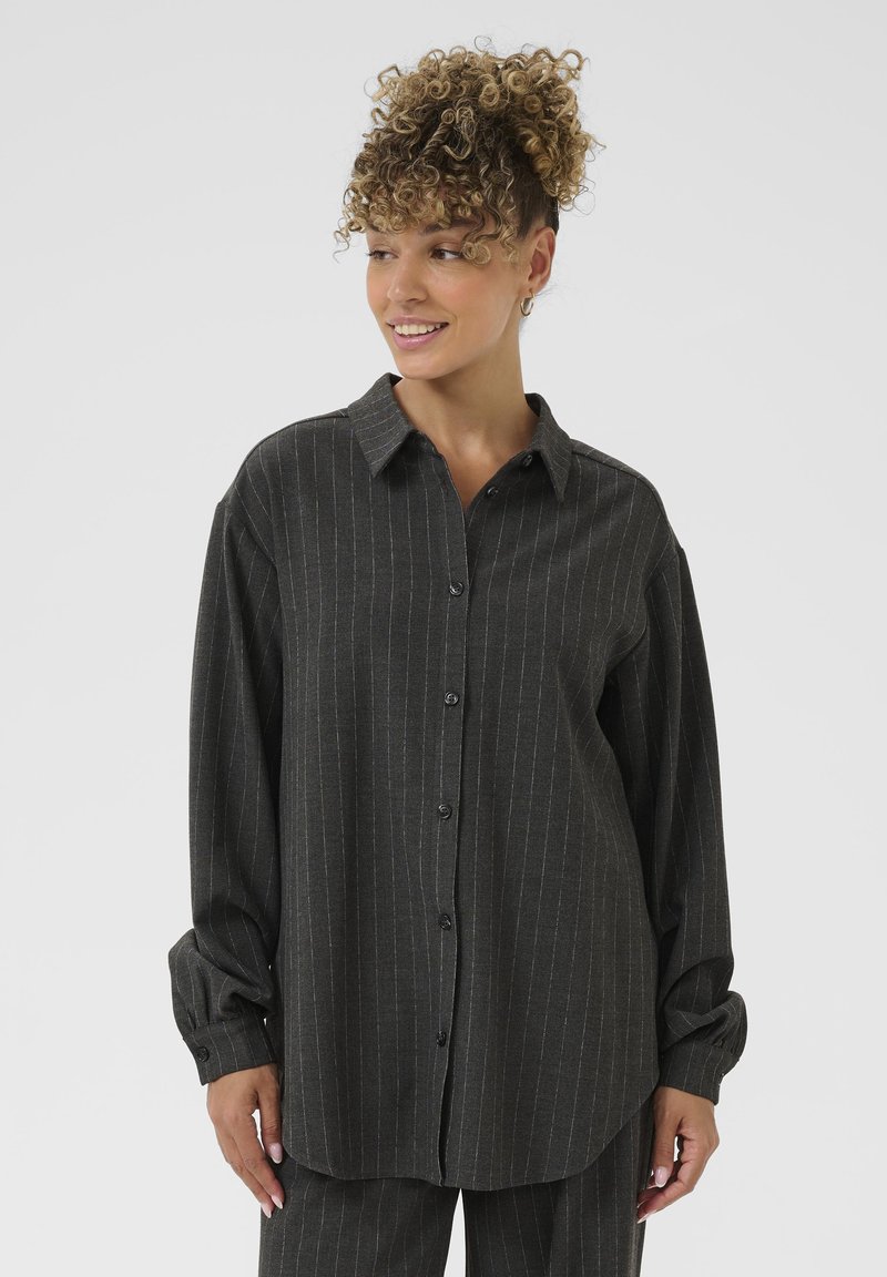Donna con capelli ricci, che indossa una camicia button-up grigio scuro a righe e pantaloni abbinati, sorridente e con lo sguardo rivolto di lato.