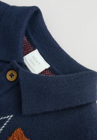 Polo en tricot bleu marine avec un col côtelé et un bouton marron. Présente un intérieur à carreaux rouges et des motifs brodés blancs, orange et bleus.