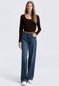 Top a maniche lunghe bordeaux a coste con dettaglio di bottoni, abbinato a jeans di denim blu larghi e cintura beige, completato da sneakers bianche.