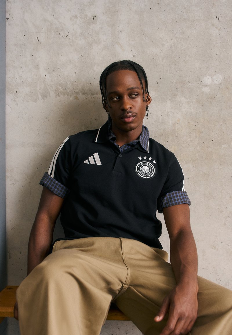 Mladý muž s copánky, oblečený v černém sportovním polo tričku a béžových kalhotách, sedí na dřevěné lavici u betonové zdi.