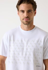 Biała koszulka wykonana z bawełny, z subtelnym wzorem w delikatne diamenty w jasnoszarym kolorze, z logo "GUESS" wydrukowanym w prawym dolnym rogu.