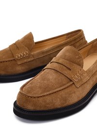 Mocassins en suède marron avec un profil bas, un détail de couture et une semelle en caoutchouc noire. Présentent une fente pour une pièce sur le dessus.